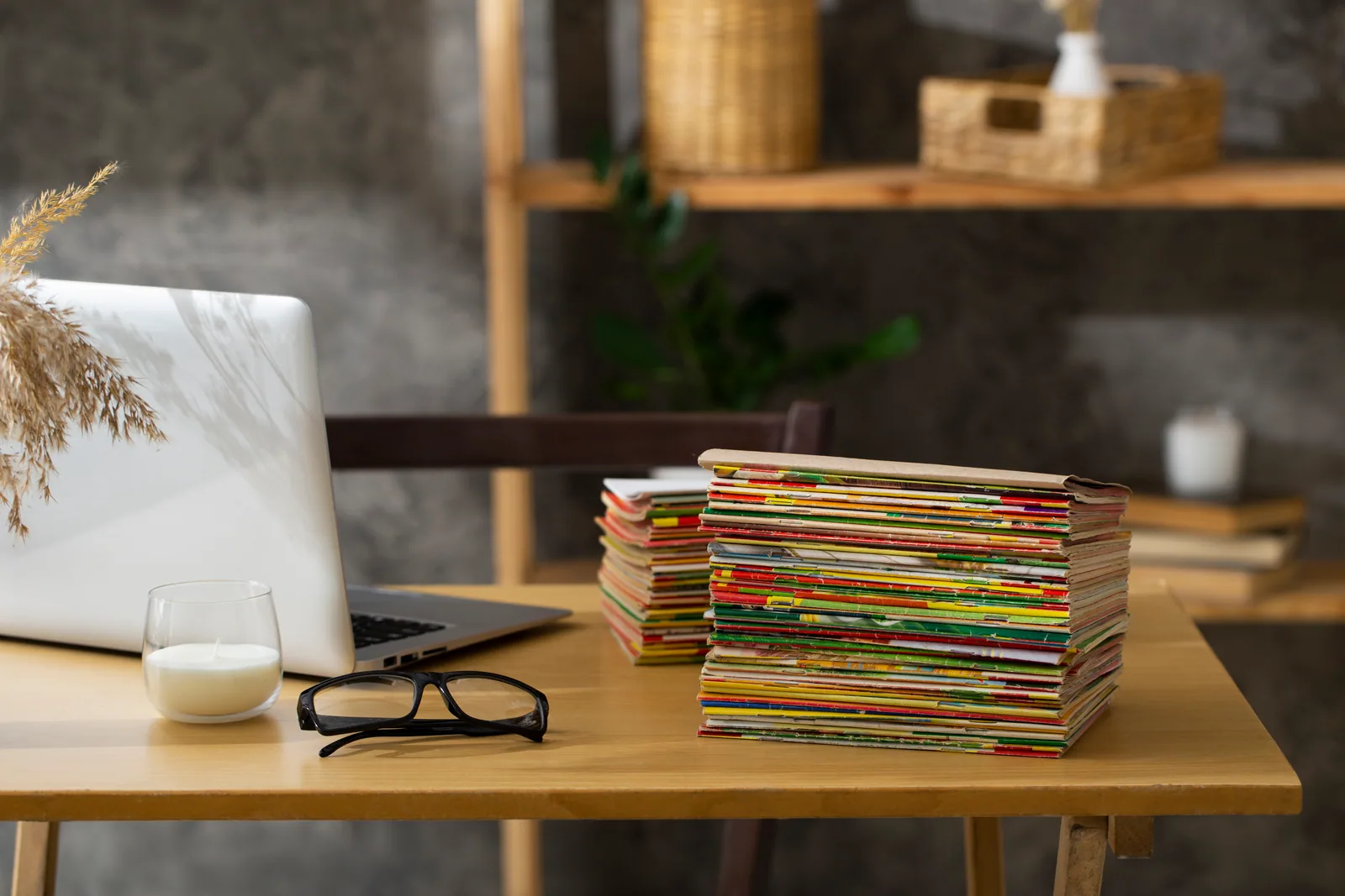 Tall stack of papers beside a laptop and reading glasses on a wooden desk