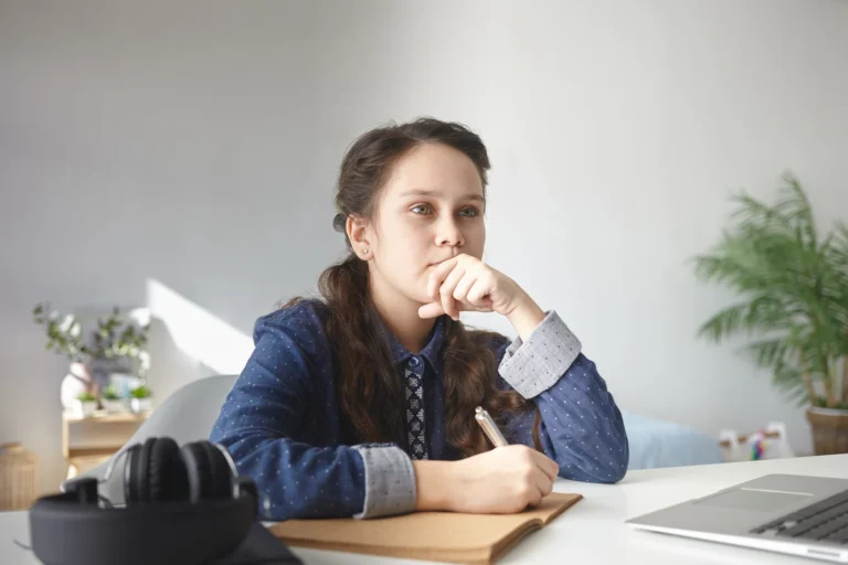 High school student thinking over an essay at a home desk with a laptop and notebook